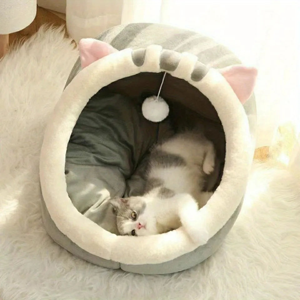 A cozy covered cat bed in gray with a cat resting inside, featuring soft plush material and a hanging pom-pom toy.