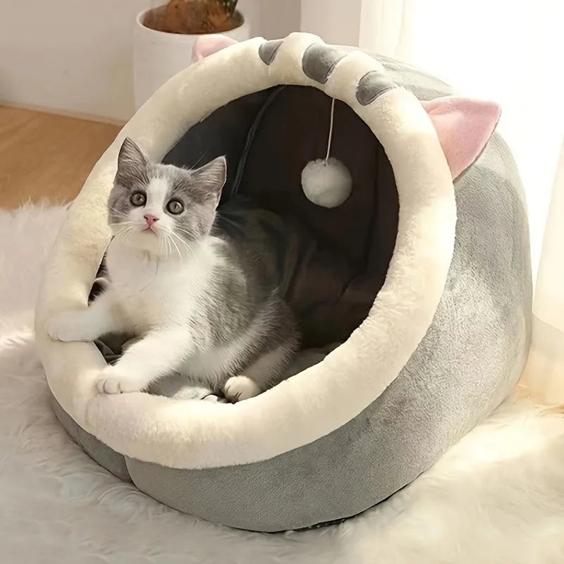 A plush gray enclosed cat bed with a cute cat resting inside and a dangling toy ball for entertainment.