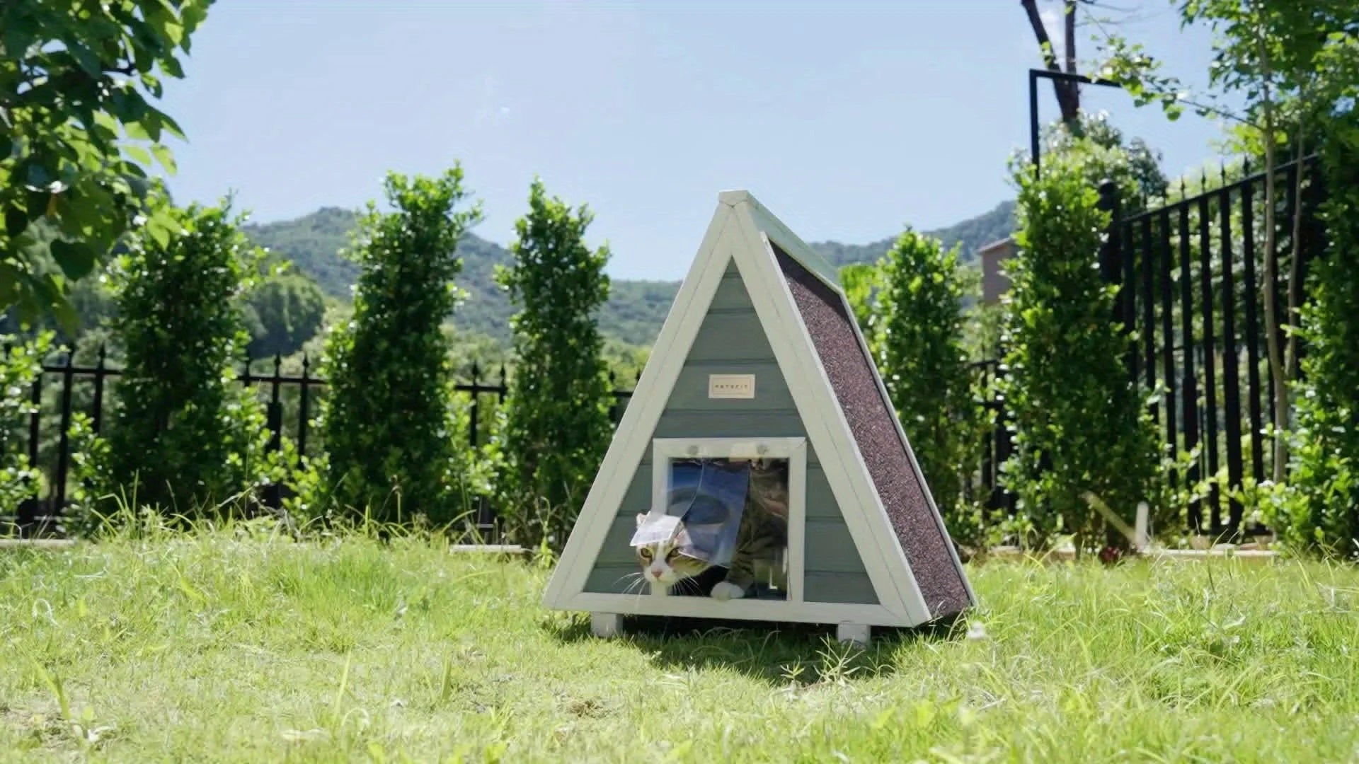 A small outdoor cat house with an A-frame design placed in a lush garden setting, featuring a cat peeking out of the entrance